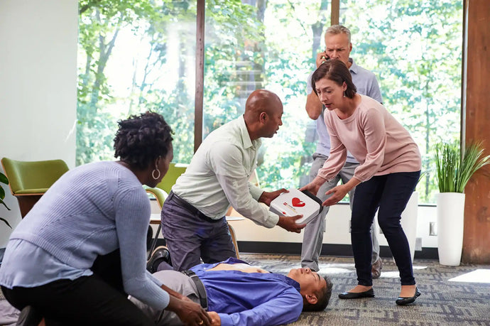 A woman passes a man a Stryker AED while a man in the background is calling an ambulance. 